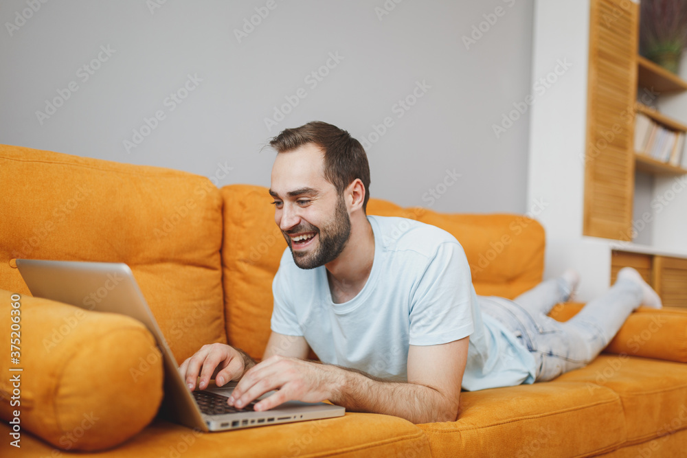 Smiling cheerful handsome attractive young bearded man 20s wearing casual blue t-shirt working on laptop pc computer lying on couch resting relaxing spending time in living room at home. Smiling cheerful handsome attractive young bearded man 20s wearing casual blue t-shirt working on laptop pc computer lying on couch resting relaxing spending time in living room at home.