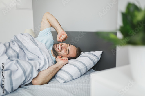 Smiling attractive young bearded man wearing basic blue t-shirt sleeping waking up stretching hands lying in bed with striped sheet pillow blanket resting relaxing spending time in bedroom at home. photo