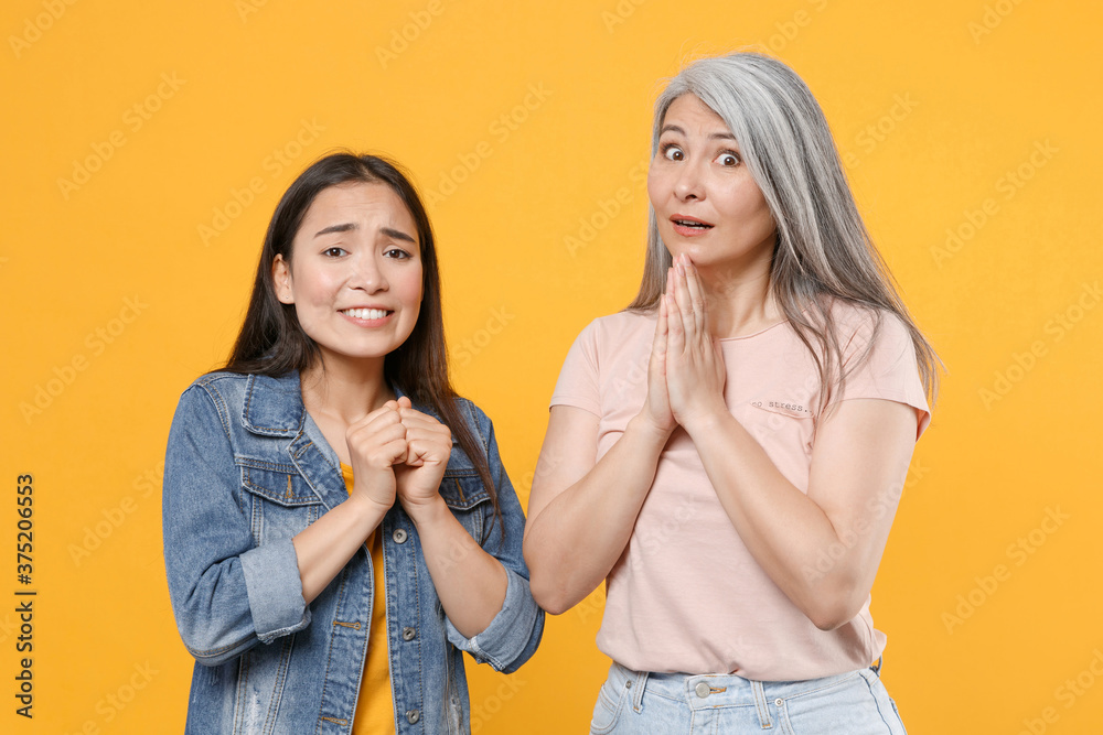 Pleading shocked family asian women girls gray-haired mother and brunette daughter in casual clothes posing holding hands folded in prayer isolated on yellow color wall background studio portrait. Pleading shocked family asian women girls gray-haired mother and brunette daughter in casual clothes posing holding hands folded in prayer isolated on yellow color wall background studio portrait.