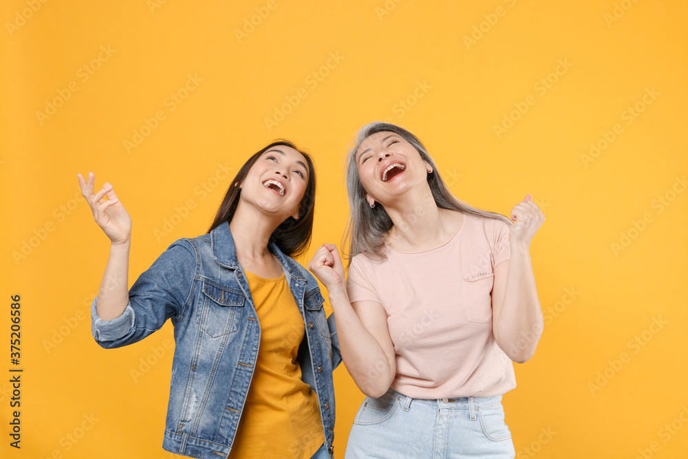 Laughing family asian women girls gray-haired mother brunette daughter in casual clothes posing dancing clenching fists rising hands looking up isolated on yellow color background studio portrait. Laughing family asian women girls gray-haired mother brunette daughter in casual clothes posing dancing clenching fists rising hands looking up isolated on yellow color background studio portrait.