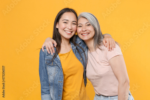 Obraz na płótnie Smiling cheerful joyful family asian female women girls gray-haired mother and brunette daughter in casual clothes posing hugging looking camera isolated on yellow color background studio portrait