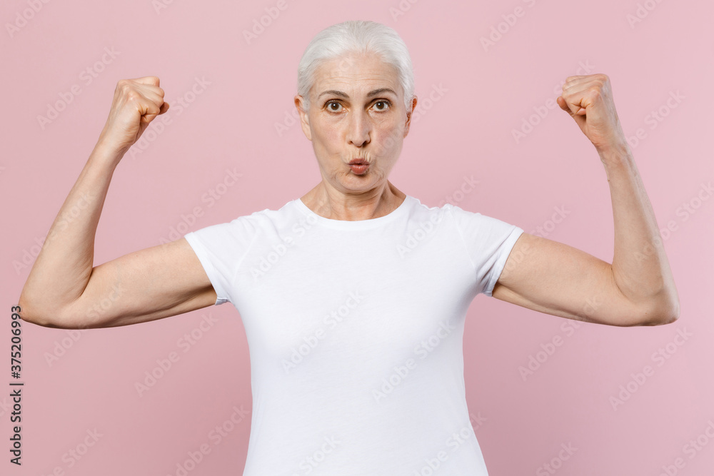 Strong shocked elderly gray-haired female woman 60s 70s wearing white design casual t-shirt posing showing biceps muscles looking camera isolated on pastel pink color wall background studio portrait. Strong shocked elderly gray-haired female woman 60s 70s wearing white design casual t-shirt posing showing biceps muscles looking camera isolated on pastel pink color wall background studio portrait.