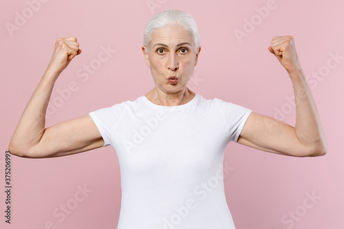 Strong shocked elderly gray-haired female woman 60s 70s wearing white design casual t-shirt posing showing biceps muscles looking camera isolated on pastel pink color wall background studio portrait.