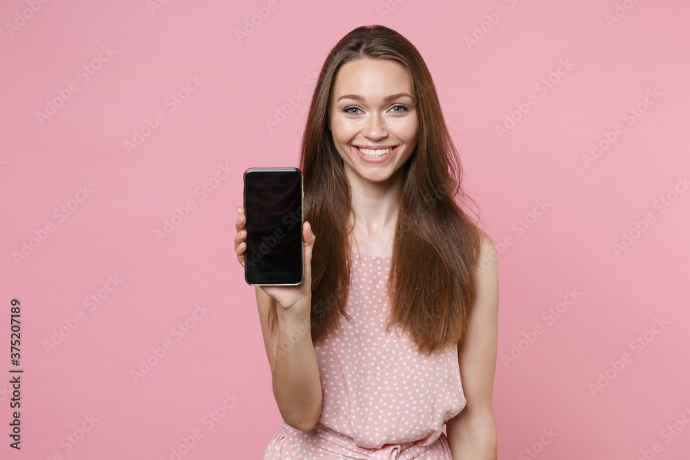 Smiling young brunette woman 20s wearing pink summer dotted dress posing hold mobile cell phone with blank empty screen mock up copy space isolated on pastel pink color background studio portrait.