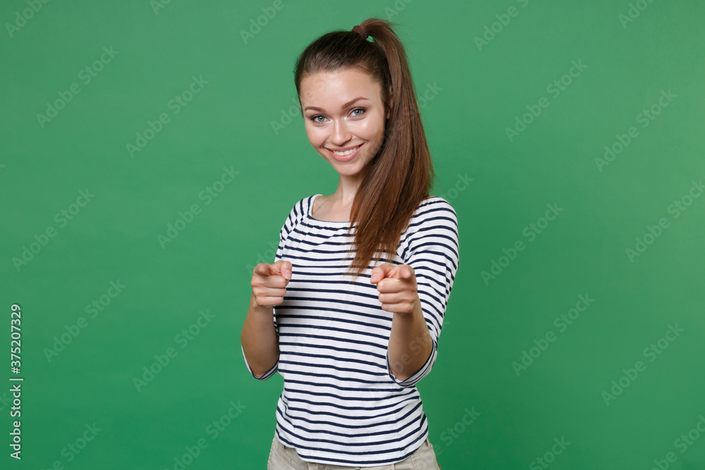 Smiling pretty beautiful attractive young brunette woman 20s wearing striped casual clothes posing standing pointing index fingers on camera isolated on green color wall background, studio portrait.