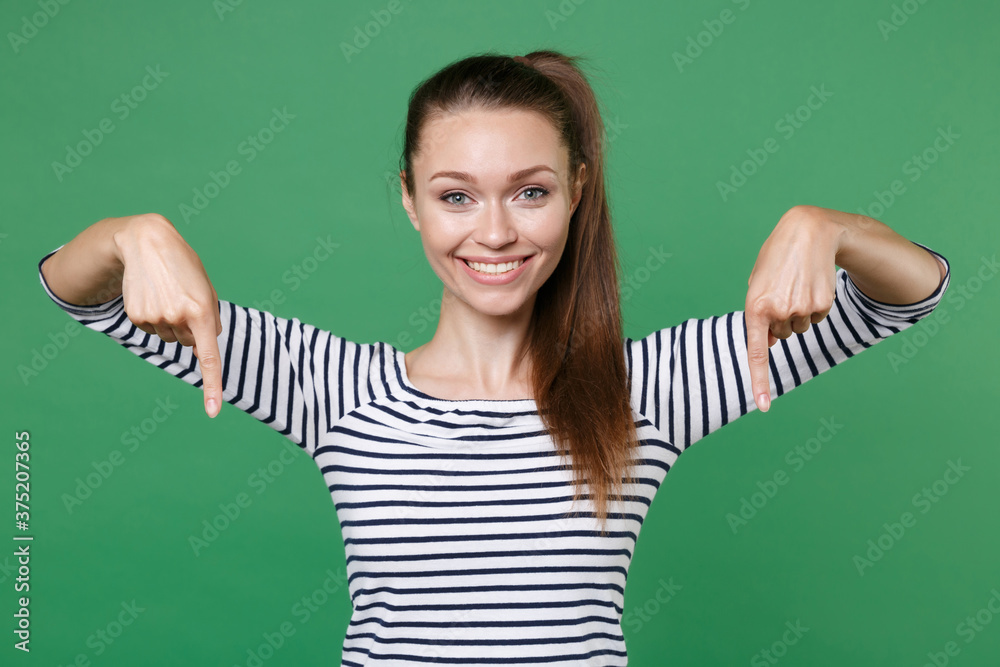 Smiling beautiful young brunette woman 20s wearing striped casual clothes posing standing pointing index fingers down on mock up copy space isolated on green color wall background studio portrait. Smiling beautiful young brunette woman 20s wearing striped casual clothes posing standing pointing index fingers down on mock up copy space isolated on green color wall background studio portrait.