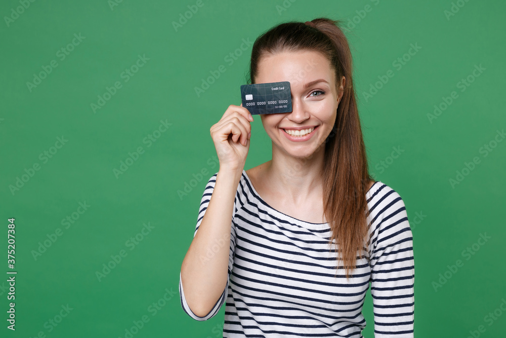 Smiling cheerful funny young brunette woman 20s wearing striped casual clothes posing holding in hand covering eye with credit bank card looking camera isolated on green background studio portrait. Smiling cheerful funny young brunette woman 20s wearing striped casual clothes posing holding in hand covering eye with credit bank card looking camera isolated on green background studio portrait.