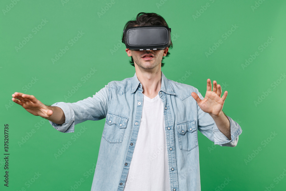 Amazed young brunet man 20s wearing casual clothes white t-shirt denim shirt watching in headset touch something like push click on button spreading hands isolated on green background studio portrait. Amazed young brunet man 20s wearing casual clothes white t-shirt denim shirt watching in headset touch something like push click on button spreading hands isolated on green background studio portrait.