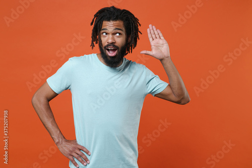 Curious young african american man guy wearing blue casual t-shirt posing isolated on orange background studio portrait. People emotions lifestyle concept. Try to hear you overhear listening intently.