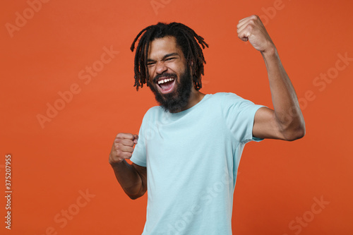 Joyful young african american man guy wearing blue casual t-shirt posing isolated on bright orange background studio portrait. People sincere emotions lifestyle concept. Clenching fists like winner.