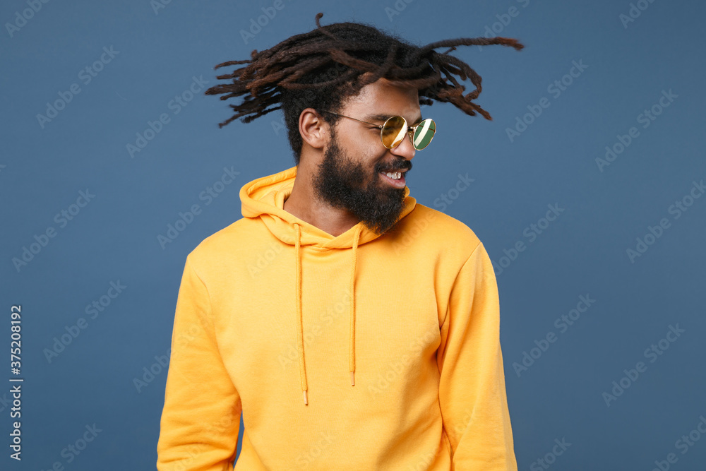 Fotka „Smiling cheerful young african american man guy wearing yellow  streetwear hoodie glasses isolated on blue wall background studio portrait.  People sincere emotions lifestyle concept. Shaking his head.“ ze služby  Stock