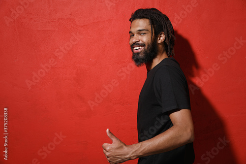 Side view of smiling young african american man guy with dreadlocks 20s wearing black casual t-shirt posing showing thumb up looking camera isolated on bright red color background studio portrait.