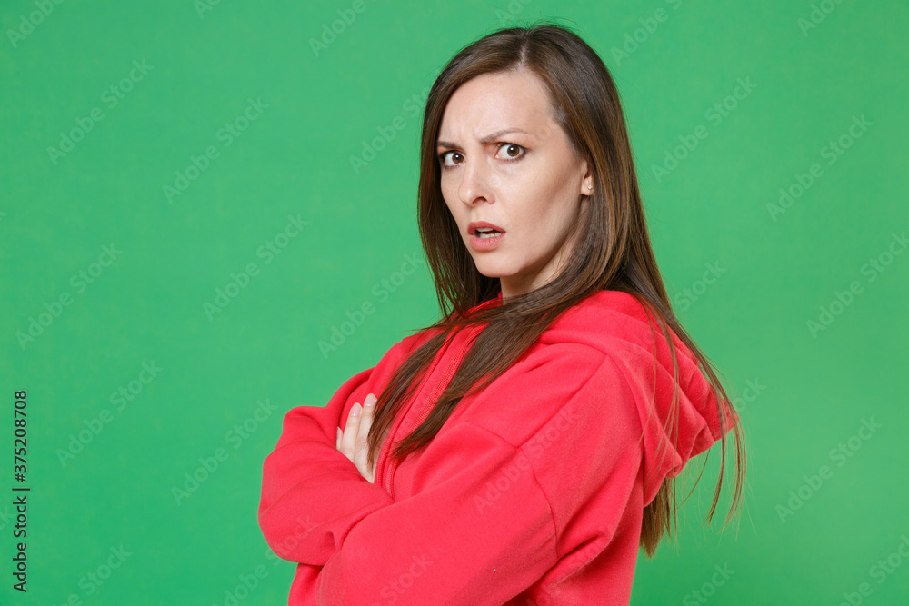 Side view of displeased perplexed young brunette woman 20s wearing bright red casual streetwear hoodie posing holding hands crossed looking camera isolated on green color background studio portrait. Side view of displeased perplexed young brunette woman 20s wearing bright red casual streetwear hoodie posing holding hands crossed looking camera isolated on green color background studio portrait.
