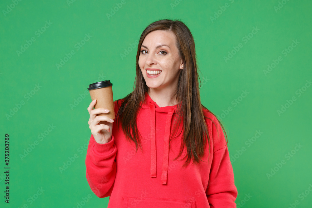 Smiling beautiful attractive young brunette woman 20s in red casual streetwear hoodie posing looking camera hold in hand paper cup of coffee or tea isolated on green color background studio portrait. Smiling beautiful attractive young brunette woman 20s in red casual streetwear hoodie posing looking camera hold in hand paper cup of coffee or tea isolated on green color background studio portrait.