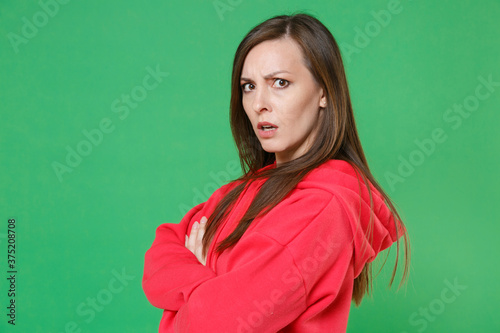 Side view of displeased perplexed young brunette woman 20s wearing bright red casual streetwear hoodie posing holding hands crossed looking camera isolated on green color background studio portrait.