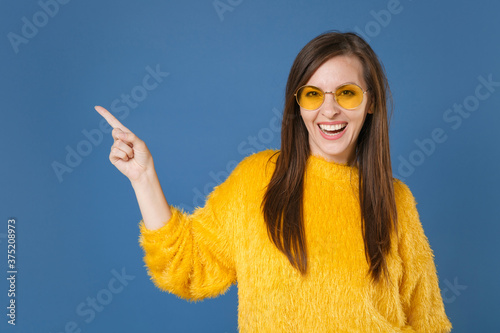 Cheerful laughing young brunette woman 20s in yellow fur sweater eyeglasses posing pointing index finger aside on mock up copy space looking camera isolated on blue color background studio portrait.
