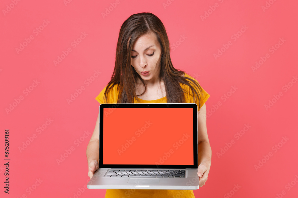 Naklejka premium Shocked amazed young brunette woman in yellow casual t-shirt posing hold looking on laptop pc computer with blank empty screen mock up copy space isolated on pink color background studio portrait.