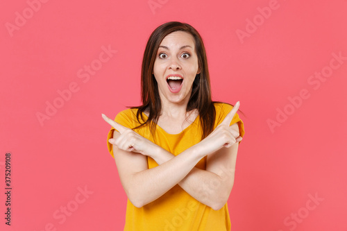 Surprised excited shocked young brunette woman 20s in yellow casual t-shirt hold hands crossed pointing index fingers aside on mock up copy space isolated on pink color background studio portrait.