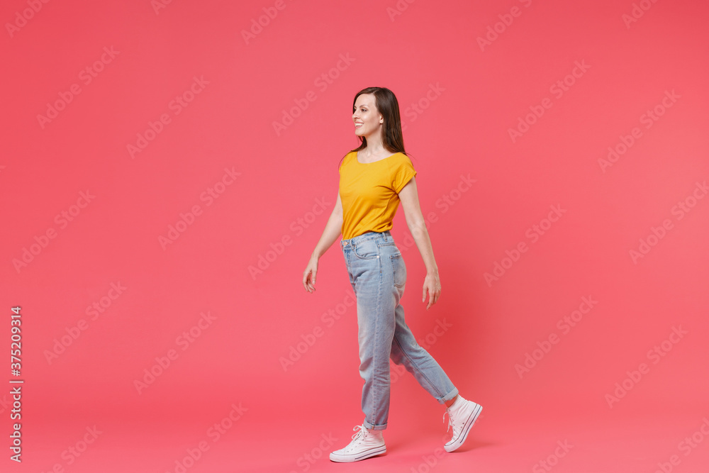 Full length portrait side view of smiling beautiful attractive young brunette woman 20s wearing yellow casual t-shirt posing walking going looking aside isolated on pink color wall background studio. Full length portrait side view of smiling beautiful attractive young brunette woman 20s wearing yellow casual t-shirt posing walking going looking aside isolated on pink color wall background studio.