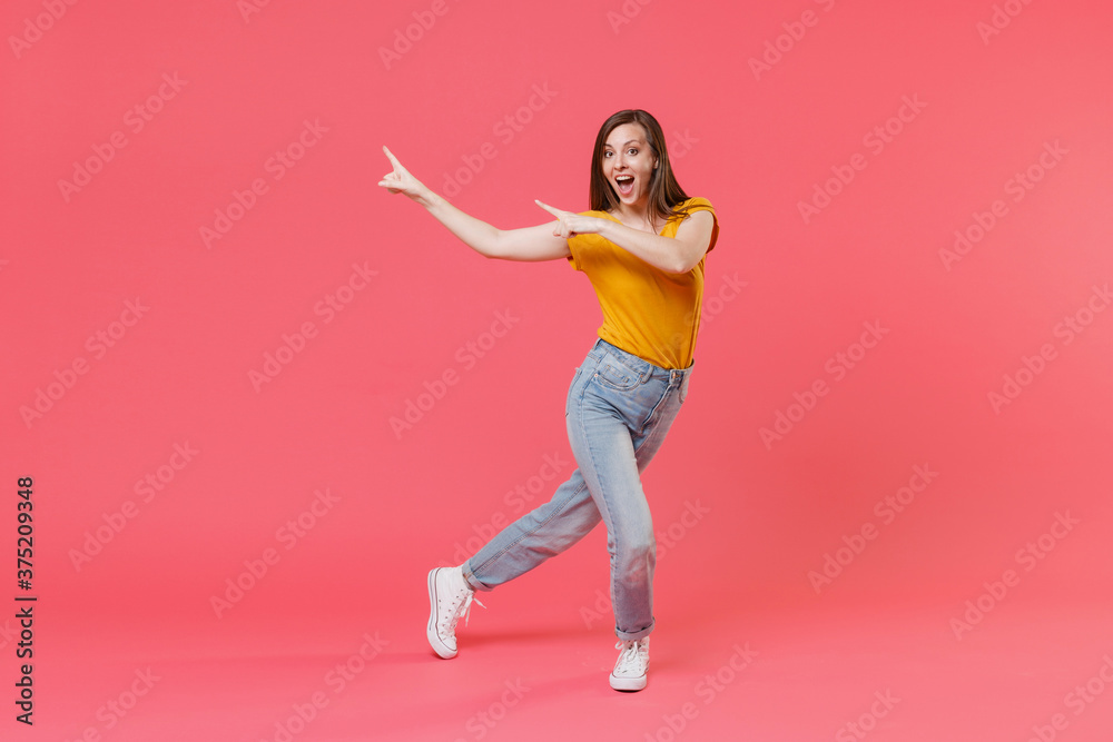 Full length portrait of excited young brunette woman 20s in yellow casual t-shirt pointing index fingers aside on mock up copy space keeping mouth open isolated on pink color wall background studio.