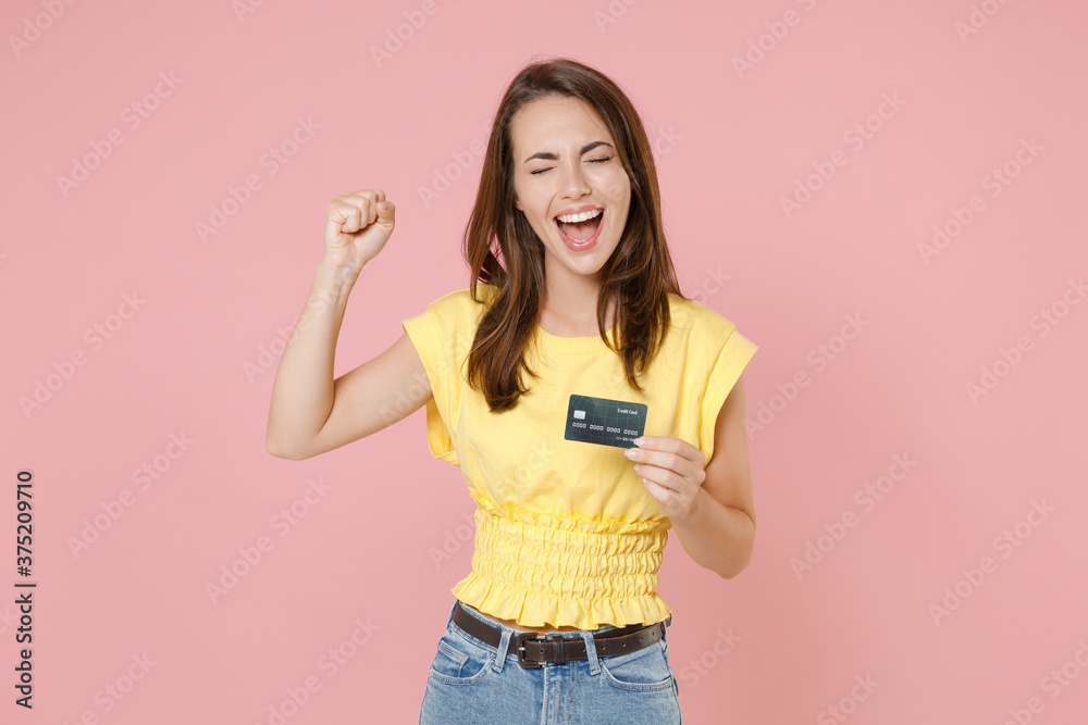 Happy joyful young brunette woman in yellow casual t-shirt posing standing holding credit bank card doing winner gesture keeping eyes closed isolated on pastel pink wall background studio portrait. Happy joyful young brunette woman in yellow casual t-shirt posing standing holding credit bank card doing winner gesture keeping eyes closed isolated on pastel pink wall background studio portrait.