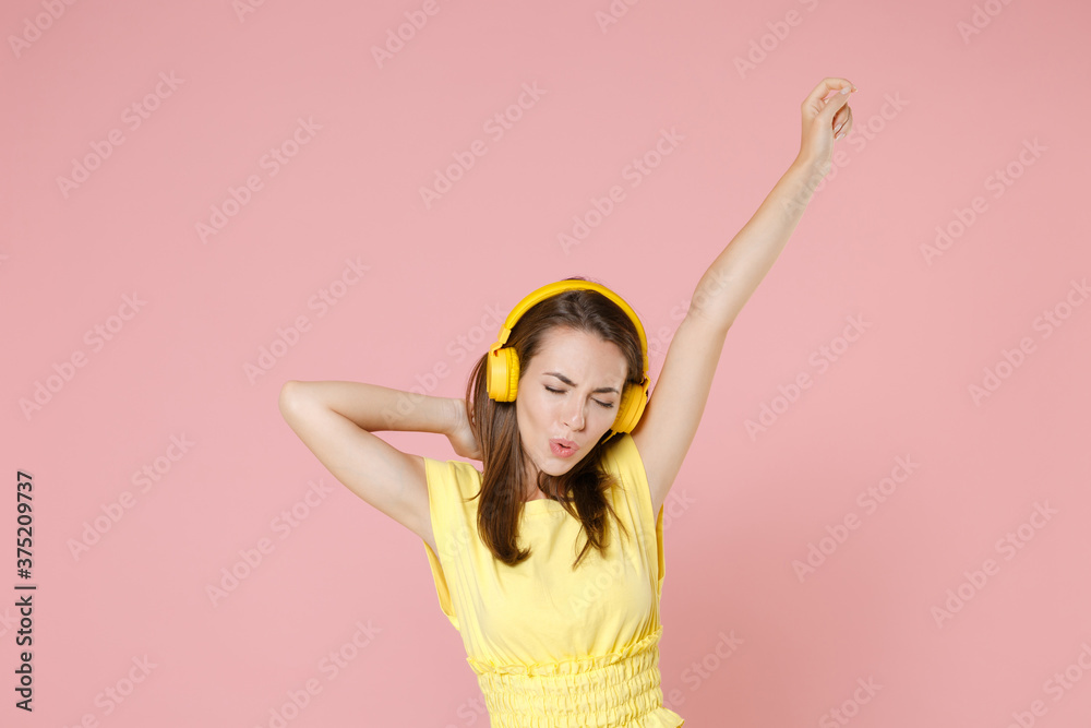 Cheerful young brunette woman 20s in yellow casual t-shirt posing listening music with headphones dancing rising hands keeping eyes closed isolated on pastel pink color background studio portrait. Cheerful young brunette woman 20s in yellow casual t-shirt posing listening music with headphones dancing rising hands keeping eyes closed isolated on pastel pink color background studio portrait.