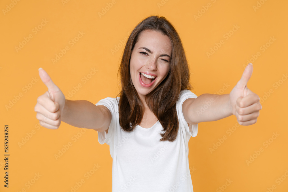 Cheerful blinking beautiful attractive young woman 20s wearing white blank empty design casual t-shirt posing standing showing thumbs up isolated on yellow color wall background studio portrait. Cheerful blinking beautiful attractive young woman 20s wearing white blank empty design casual t-shirt posing standing showing thumbs up isolated on yellow color wall background studio portrait.