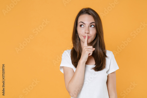 Secret young woman 20s in white blank empty design casual t-shirt posing saying hush be quiet with finger on lips shhh gesture looking aside up isolated on yellow color background studio portrait.