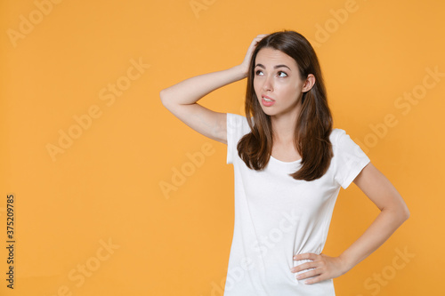 Preoccupied puzzled concerned young brunette woman 20s in white blank empty design casual t-shirt posing standing put hand on head looking aside up isolated on yellow color background studio portrait.
