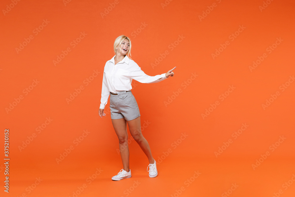 Full length portrait of smiling cheerful funny young blonde female business woman girl 20s in white shirt posing pointing index finger aside isolated on bright orange color wall background studio. Full length portrait of smiling cheerful funny young blonde female business woman girl 20s in white shirt posing pointing index finger aside isolated on bright orange color wall background studio.