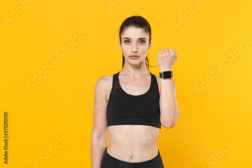 Beautiful young fitness sporty woman 20s wearing black sportswear posing working out training wearing smart watch on hand looking camera isolated on bright yellow color background studio portrait.