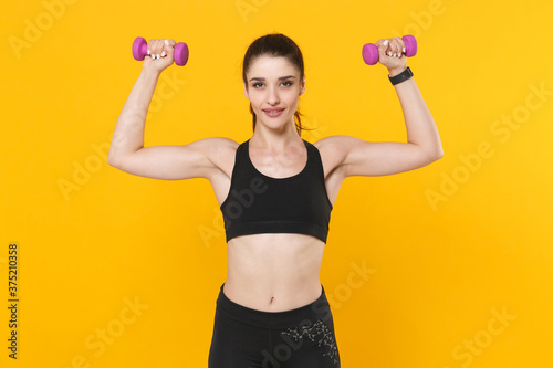 Pretty young fitness sporty woman 20s in black sportswear posing training working out doing exercise with dumbbells spreading hands looking camera isolated on yellow color background studio portrait.