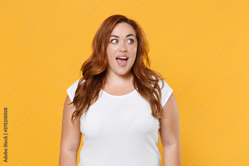 Excited young redhead plus size body positive female woman girl 20s in white blank design casual t-shirt posing keeping mouth open looking aside isolated on yellow color background studio portrait. Excited young redhead plus size body positive female woman girl 20s in white blank design casual t-shirt posing keeping mouth open looking aside isolated on yellow color background studio portrait.