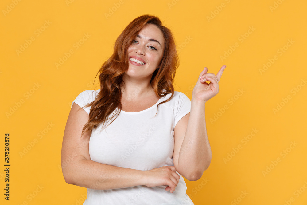 Smiling young redhead plus size body positive female woman girl 20s in white casual t-shirt posing pointing index finger up on mock up copy space isolated on yellow color background studio portrait. Smiling young redhead plus size body positive female woman girl 20s in white casual t-shirt posing pointing index finger up on mock up copy space isolated on yellow color background studio portrait.