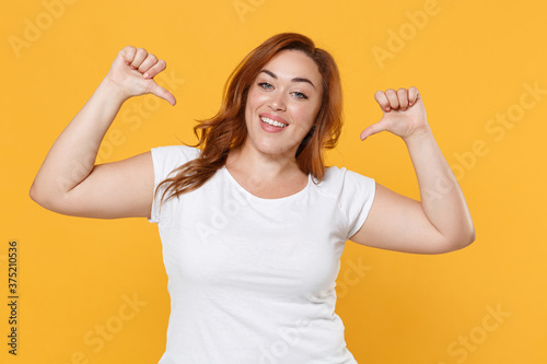 Smiling cheerful young redhead plus size body positive female woman girl 20s in white blank design casual t-shirt posing pointing thumbs on herself isolated on yellow color background studio portrait.