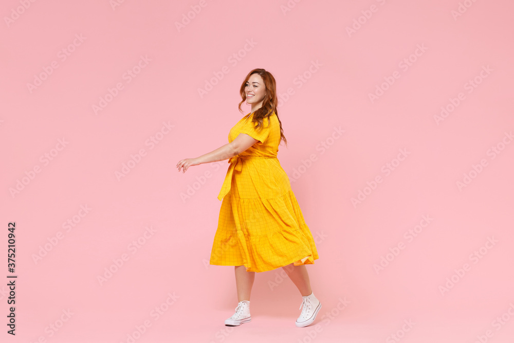 Full length portrait smiling attractive young redhead plus size body positive female woman girl 20s in yellow dress posing walking going looking aside isolated on pastel pink color background studio. Full length portrait smiling attractive young redhead plus size body positive female woman girl 20s in yellow dress posing walking going looking aside isolated on pastel pink color background studio.