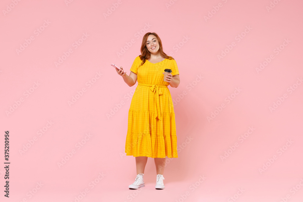 Full length portrait smiling young redhead plus size body positive female woman 20s in yellow dress hold cup of coffee tea mobile phone looking camera isolated on pastel pink color background studio. Full length portrait smiling young redhead plus size body positive female woman 20s in yellow dress hold cup of coffee tea mobile phone looking camera isolated on pastel pink color background studio.