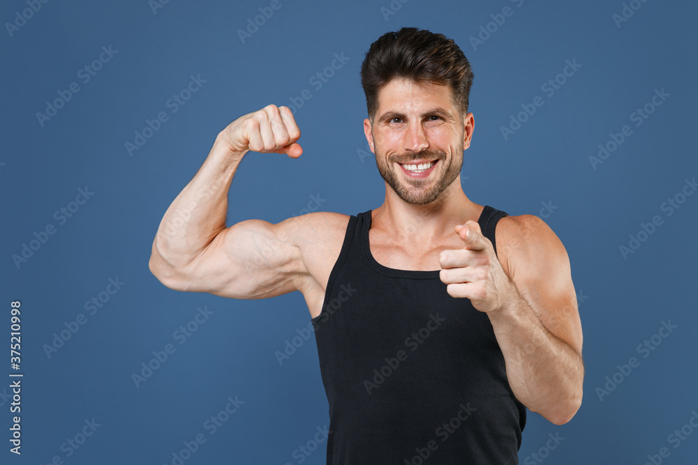 Smiling young bearded fitness sporty guy sportsman in black undershirt isolated on blue background. Workout sport motivation lifestyle concept. Showing biceps muscles pointing index finger on camera. Smiling young bearded fitness sporty guy sportsman in black undershirt isolated on blue background. Workout sport motivation lifestyle concept. Showing biceps muscles pointing index finger on camera.