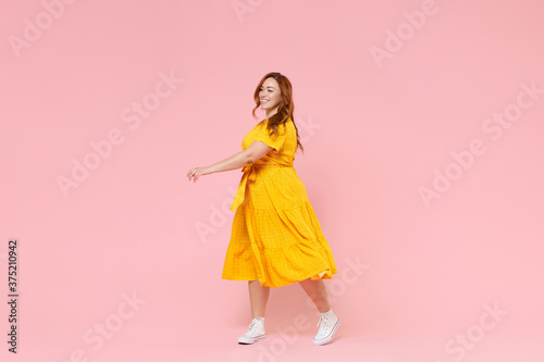 Full length portrait smiling attractive young redhead plus size body positive female woman girl 20s in yellow dress posing walking going looking aside isolated on pastel pink color background studio.