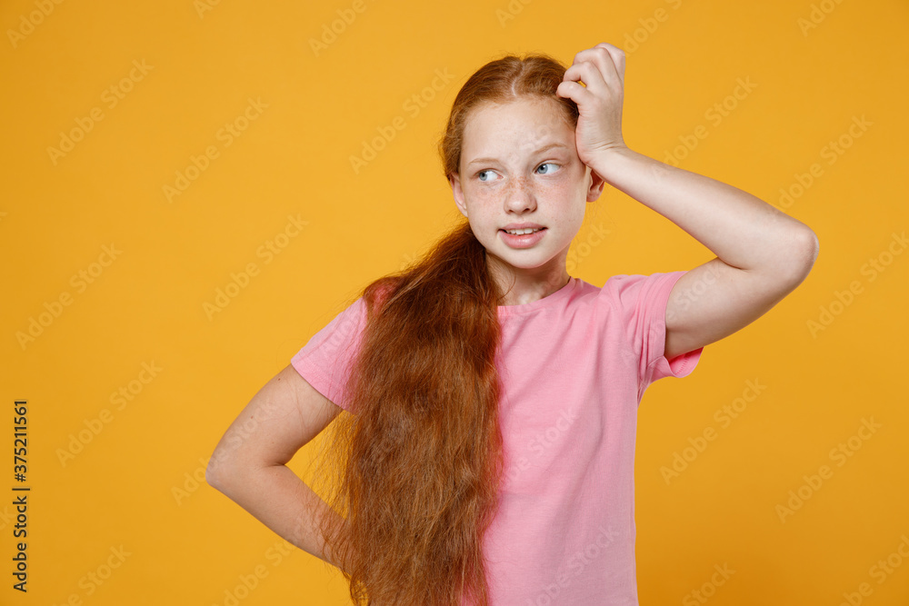 Pensive puzzled little ginger redhead kid girl 12-13 years old wearing pink casual t-shirt posing put hand on head looking aside isolated on bright yellow color background children studio portrait. Pensive puzzled little ginger redhead kid girl 12-13 years old wearing pink casual t-shirt posing put hand on head looking aside isolated on bright yellow color background children studio portrait.