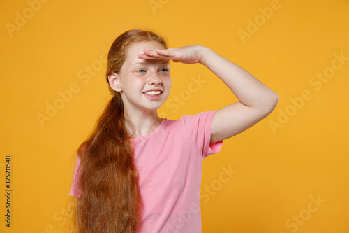Smiling little ginger redhead kid girl 12-13 years old in pink casual t-shirt posing holding hand at forehead looking far away distance isolated on yellow color background children studio portrait.