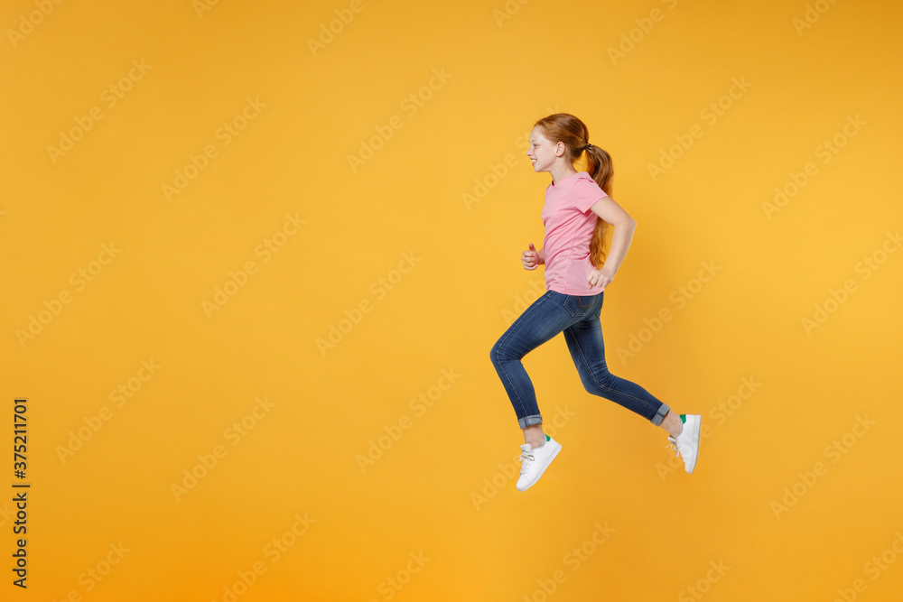 Full length children studio portrait side view of smiling little ginger redhead kid girl 12-13 years old wearing pink casual t-shirt posing jumping like running isolated on yellow color background.