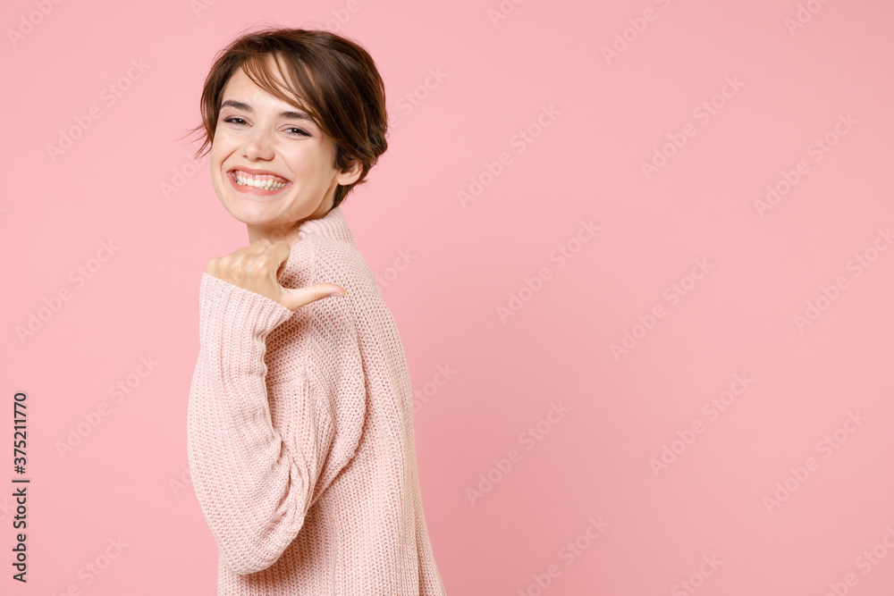 Side view of smiling young brunette woman girl in knitted casual sweater posing isolated on pastel pink background studio portrait. People lifestyle concept. Mock up copy space. Pointing thumb aside. Side view of smiling young brunette woman girl in knitted casual sweater posing isolated on pastel pink background studio portrait. People lifestyle concept. Mock up copy space. Pointing thumb aside.