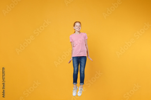 Full length children studio portrait of smiling little ginger redhead kid girl 12-13 years old wearing pink casual t-shirt posing jumping having fun isolated on bright yellow color wall background.