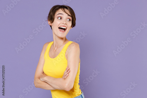 Side view of excited young brunette woman girl in yellow casual tank top posing isolated on pastel violet background studio portrait. People lifestyle concept. Holding hands crossed, looking aside.