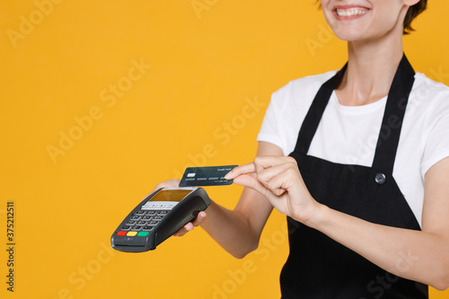 Cropped image young woman barista bartender barman employee in apron hold wireless modern bank payment terminal to process acquire credit card payments isolated on yellow background studio portrait.