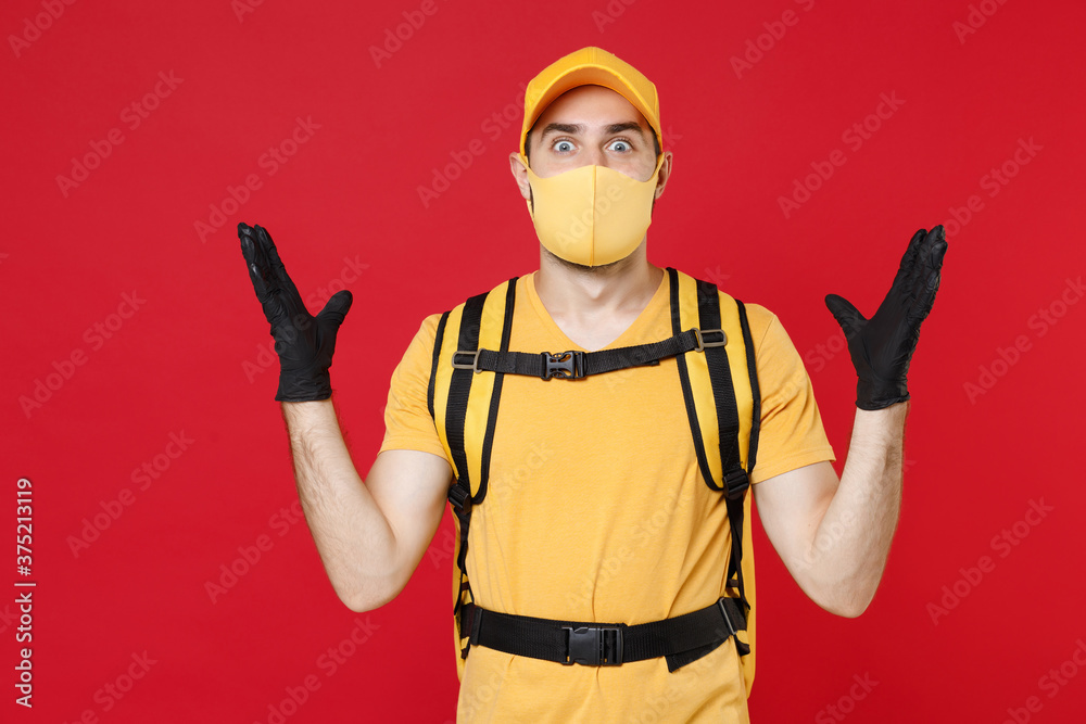 Delivery employee man in yellow cap face mask gloves t-shirt uniform thermal bag backpack with food work courier service during quarantine coronavirus covid-19 virus isolated on red background studio.