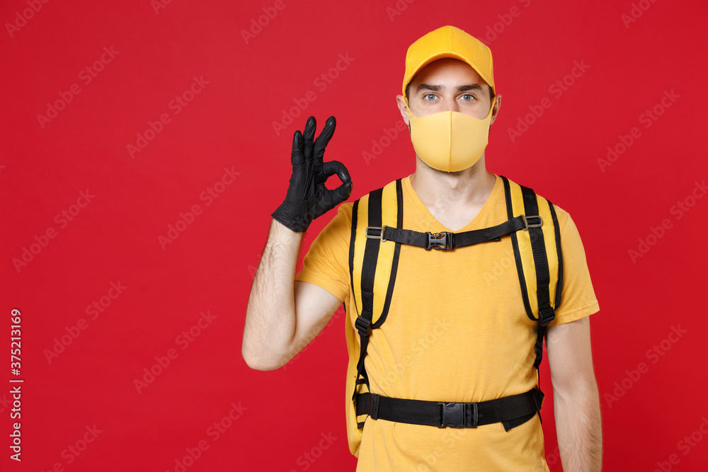 Delivery employee man in yellow cap face mask gloves t-shirt uniform thermal bag backpack with food work courier service during quarantine coronavirus covid-19 virus isolated on red background studio. Delivery employee man in yellow cap face mask gloves t-shirt uniform thermal bag backpack with food work courier service during quarantine coronavirus covid-19 virus isolated on red background studio.