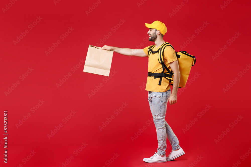 Full length side view step delivery employee man male in yellow cap t-shirt thermal backpack hold craft paper food takeaway bag work courier service in quarantine covid-19 isolated on red background.