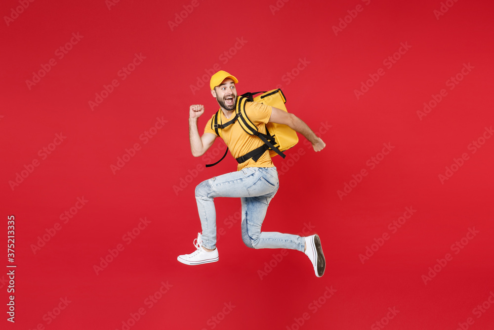 Full length delivery employee man guy male 20s in yellow cap t-shirt uniform thermal food bag backpack work courier service during quarantine covid-19 virus jumping isolated on red background studio.